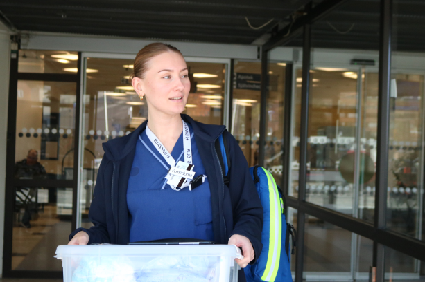 Nurse in blue scrubs carrying medical equipment outside a healthcare facility, prepared for a home visit