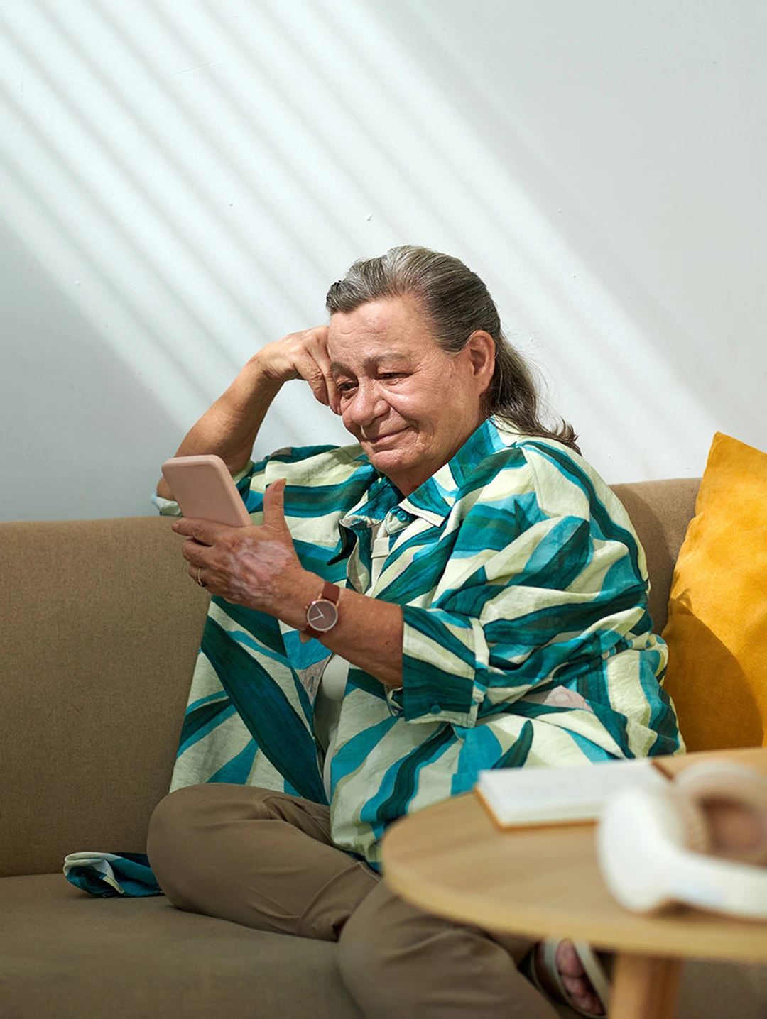 Older woman sitting on a sofa smiling while interacting with a smartphone, representing patient engagement at home